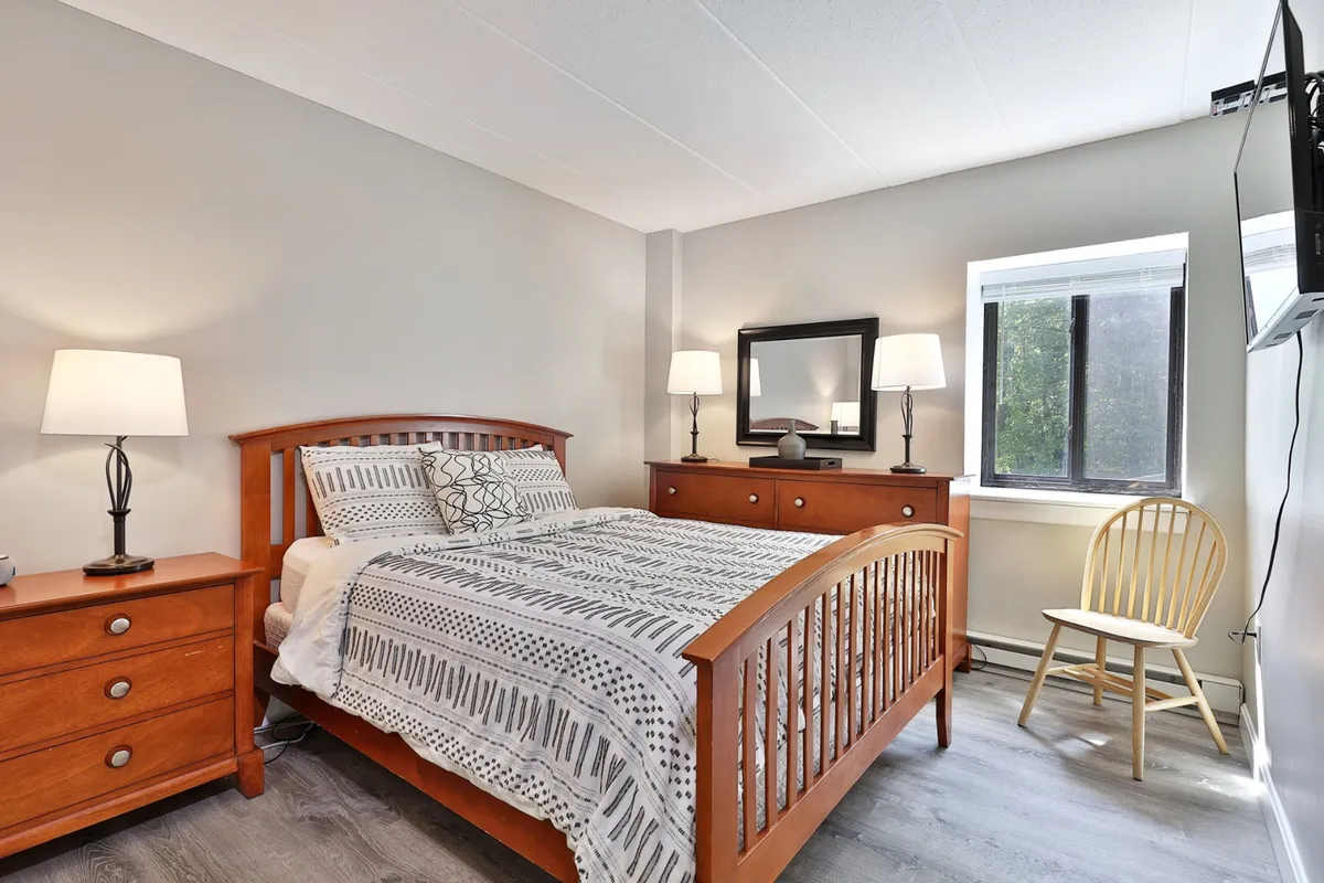 A serene bedroom featuring a plush bed with crisp white linens, a nightstand with a lamp, a mirror, and a large window allowing natural light to filter in. The space has a tranquil, uncluttered ambiance, perfect for rest and relaxation.