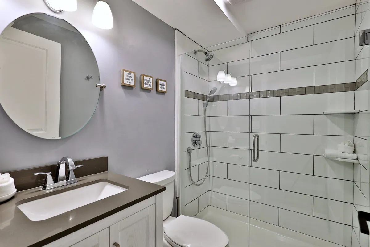 A modern bathroom featuring a circular mirror above a vanity with a sink, stylish framed artwork, and a glass shower enclosure. The space is well-lit and designed for comfort and convenience.