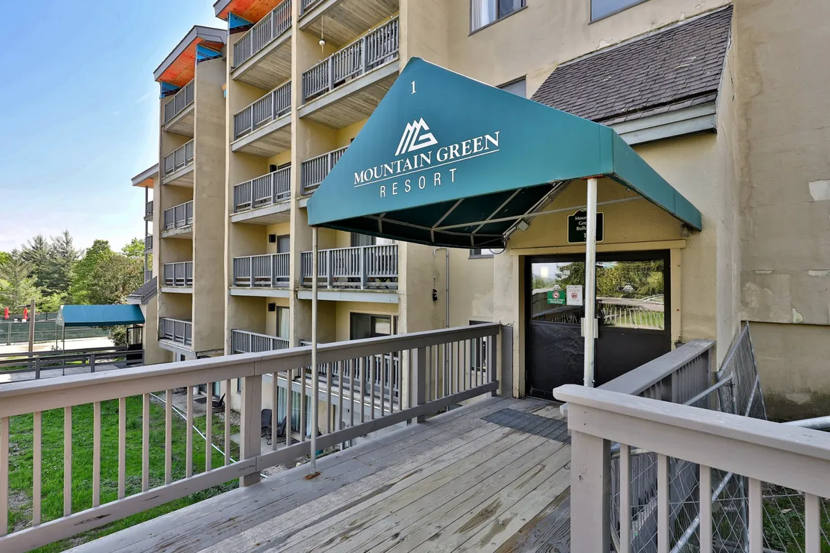 An exterior view of the building showing entrance awning, balconies, and a wooden ramp. The entry feels accessible and welcoming, indicating hassle-free parking nearby and a well-maintained exterior—matching the description of practical and resort-style perks.