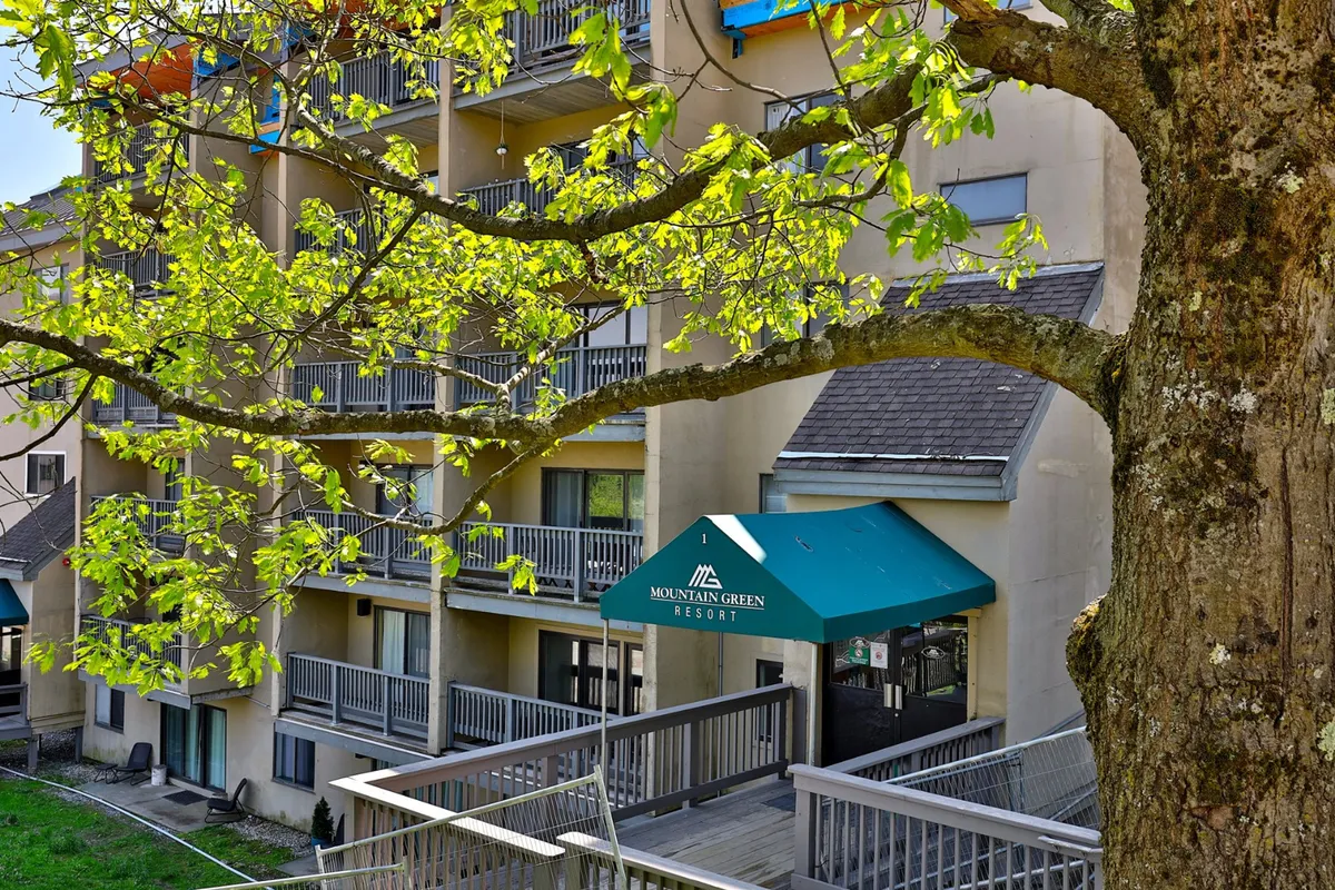 This image highlights the exterior entrance of Mountain Green Resort, featuring the welcoming canopy with the resort's sign, multiple balconies, and lush greenery, all under inviting daylight. The scene emphasizes the connection between the property and its vibrant surroundings, suggesting easy access to the local nightlife and après-ski venues.