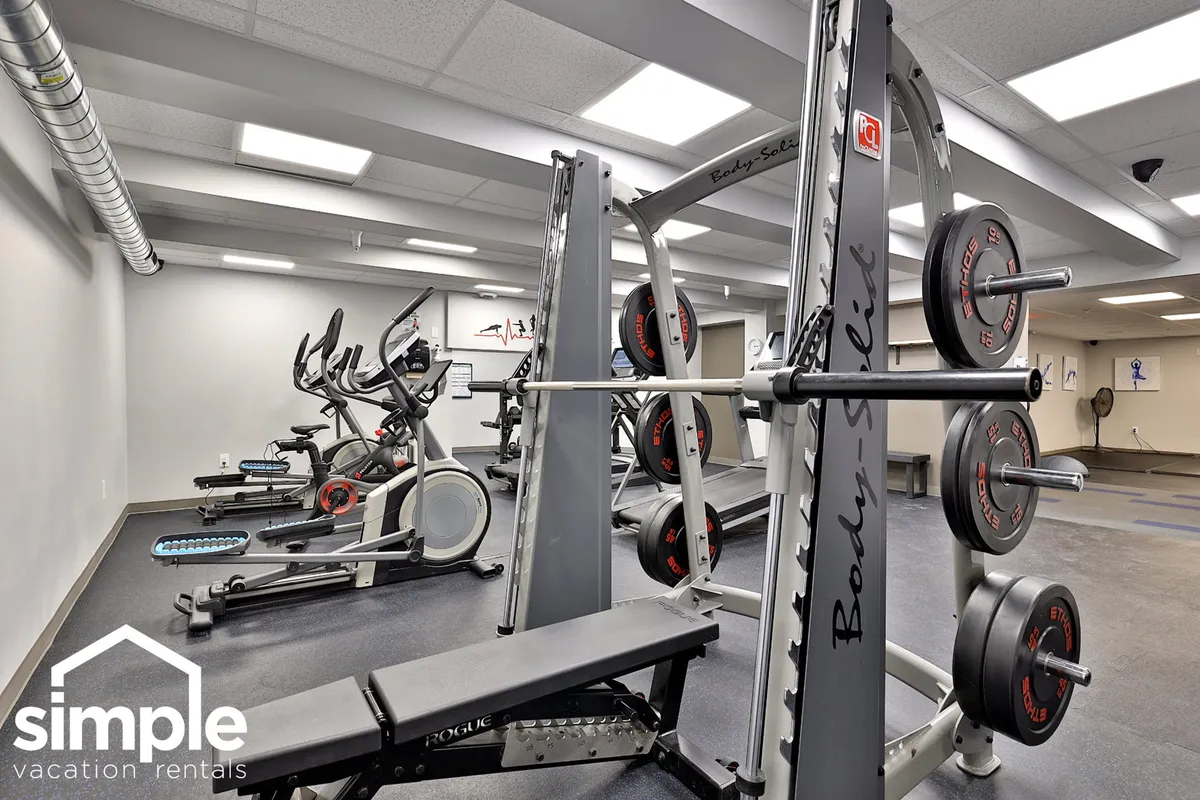 A bright fitness room featuring weightlifting equipment, exercise bikes, and mirrors. The clean modern space encourages an active lifestyle and suggests the presence of well-maintained communal amenities accessible to residents.