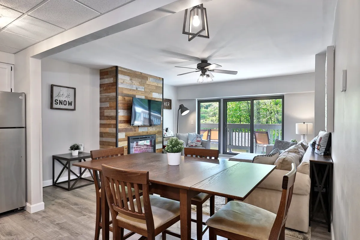 An open-plan area with a stone feature wall, a welcoming dining table set for several people, large windows letting in natural light, and a comfortable seating area adjacent to the dining space. The fireplace and TV are visible, enhancing the sense of flow between living and dining zones.