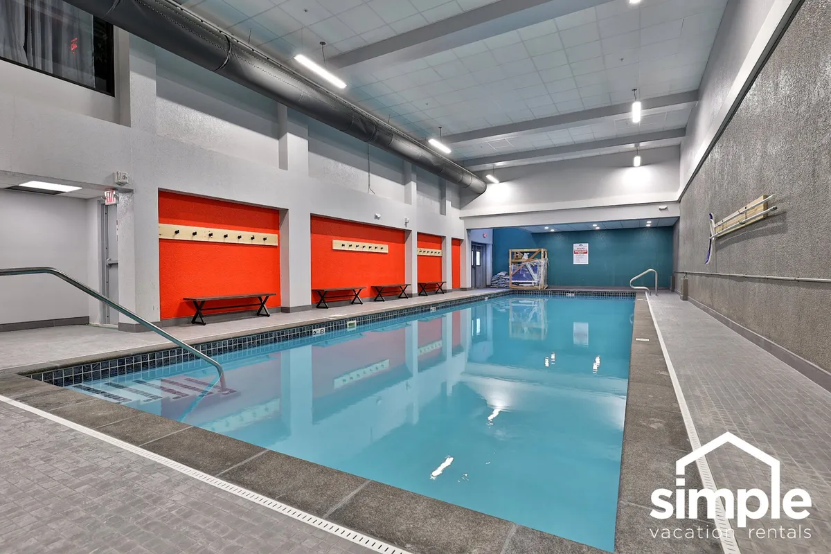 A bright indoor swimming pool area with vibrant red accent walls, benches lining the poolside, and clear, inviting water. The space is well-lit and designed for both relaxation and social gatherings.