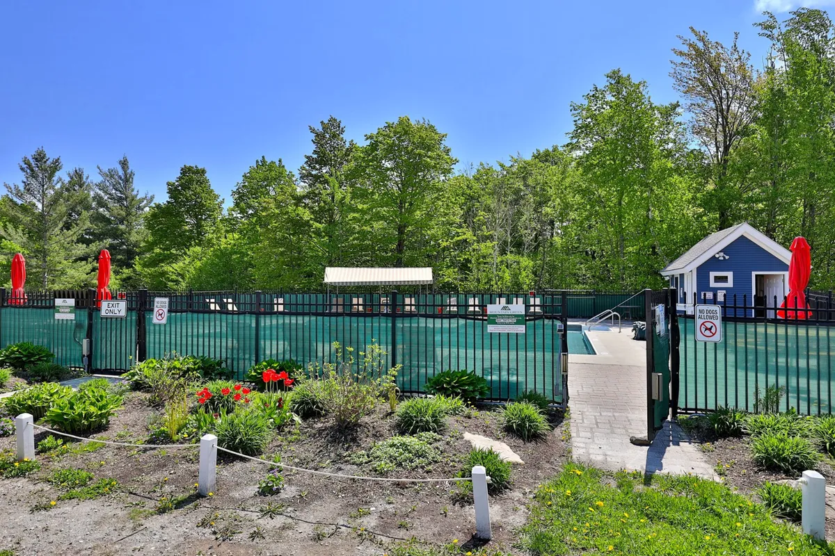 A vibrant exterior shot of the resort's heated outdoor saltwater pool surrounded by a landscaped garden, red umbrellas, and a blue building. The scene exudes summer fun, relaxation, and a sense of community. Sun loungers and poolside spaces are visible, highlighting areas perfect for families to gather, swim, and unwind during warm months.