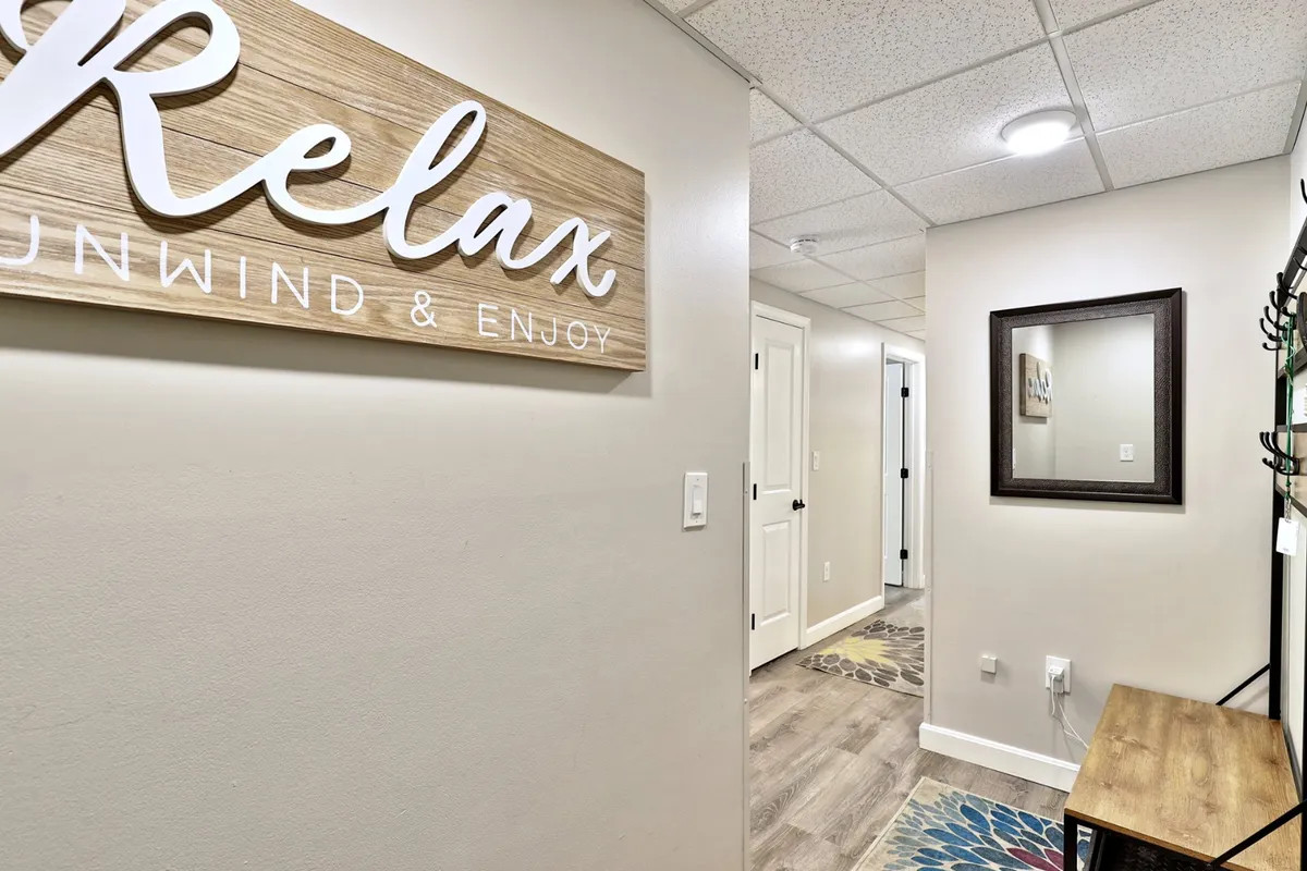 A welcoming entryway featuring a 'Relax' sign, a mirror for last checks before heading out, a sturdy wooden bench for sitting or organizing shoes, and a colorful rug adding a touch of warmth. The vibe is calm and homey, clearly presenting the first stop when coming in and out of the condo.