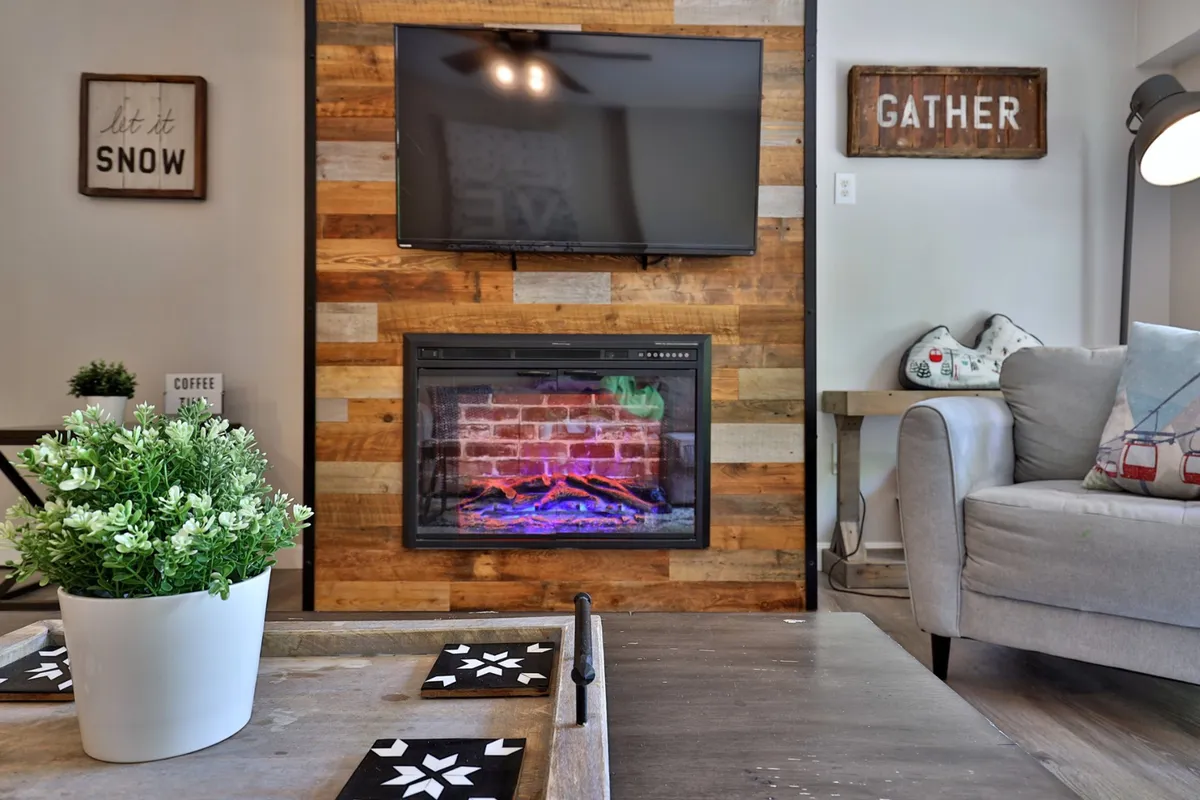 A warm and welcoming living room featuring a modern electric fireplace set into a wood-paneled wall, a sleek TV mounted above, a low coffee table with coasters, and comfortable seating arranged for gatherings. Decorative signs add a personal touch, and ample natural light enhances the inviting atmosphere.
