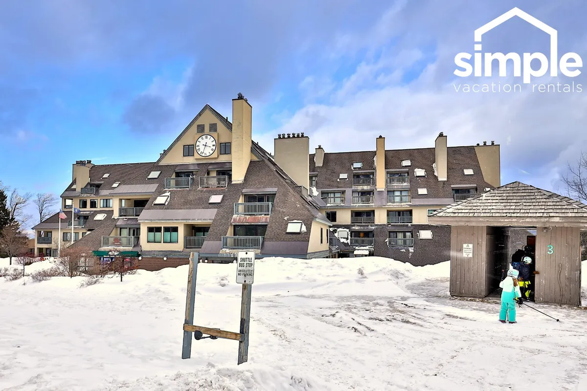 This image shows the exterior of a building at Killington Resort, with a distinctive clock tower, snow-covered ground, and people dressed in winter attire. The atmosphere is lively, with visitors heading out for winter activities like skiing and snowboarding, set against a backdrop of fresh snow and clear winter light.